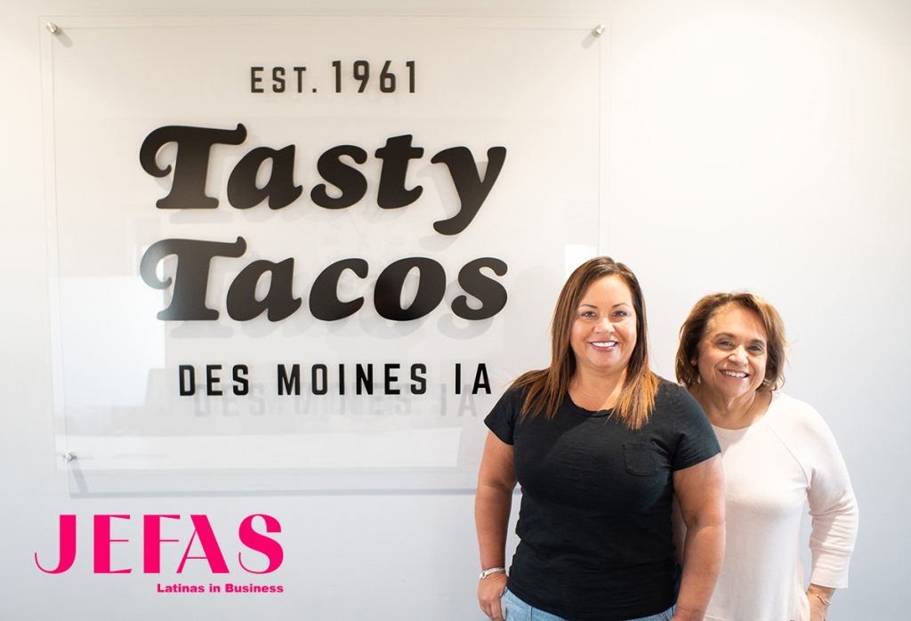 2 Women - one wearing black shirt, older woman behind her wearing white shirt, smile at camera in front of Tasty Tacos sign