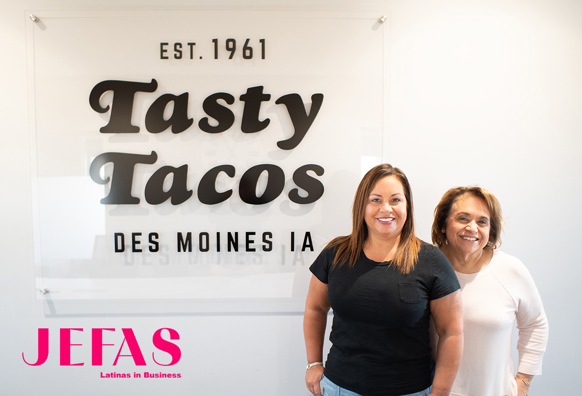 2 Women - one wearing black shirt, older woman behind her wearing white shirt, smile at camera in front of Tasty Tacos sign